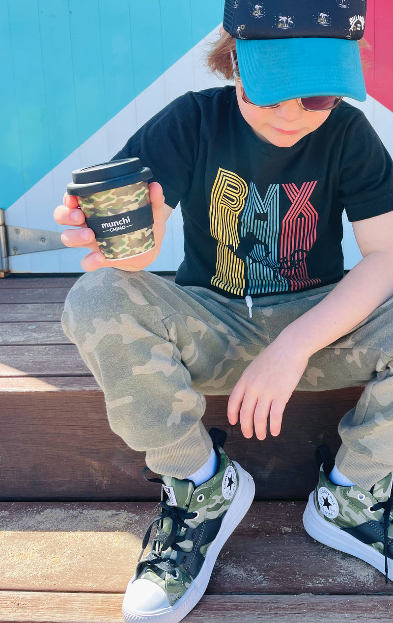 kid wearing cap and camouflage converse shoes holding camouflage babychino cup with black lid